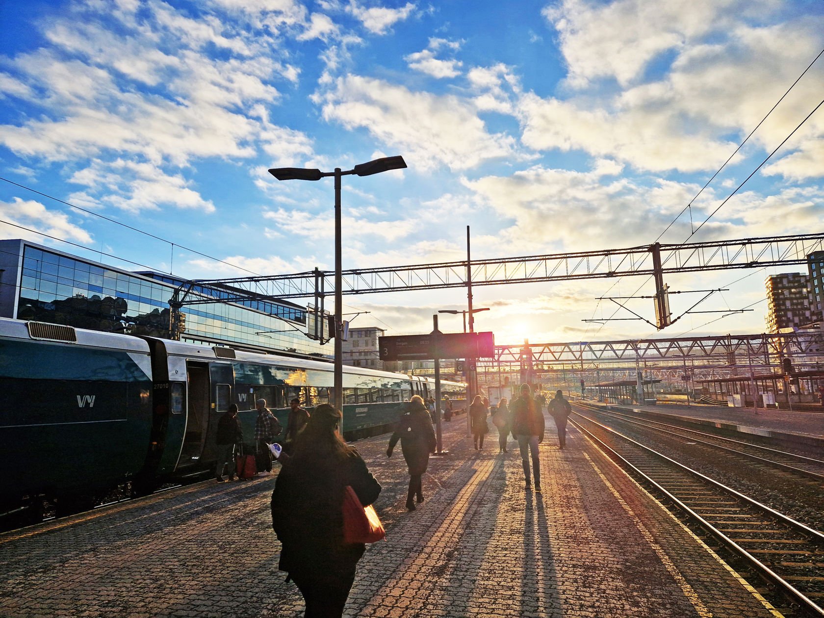 Lav sol over perrongene på Oslo S. Folk går på plattformen mot sola. Et tog står på ett av sporene.