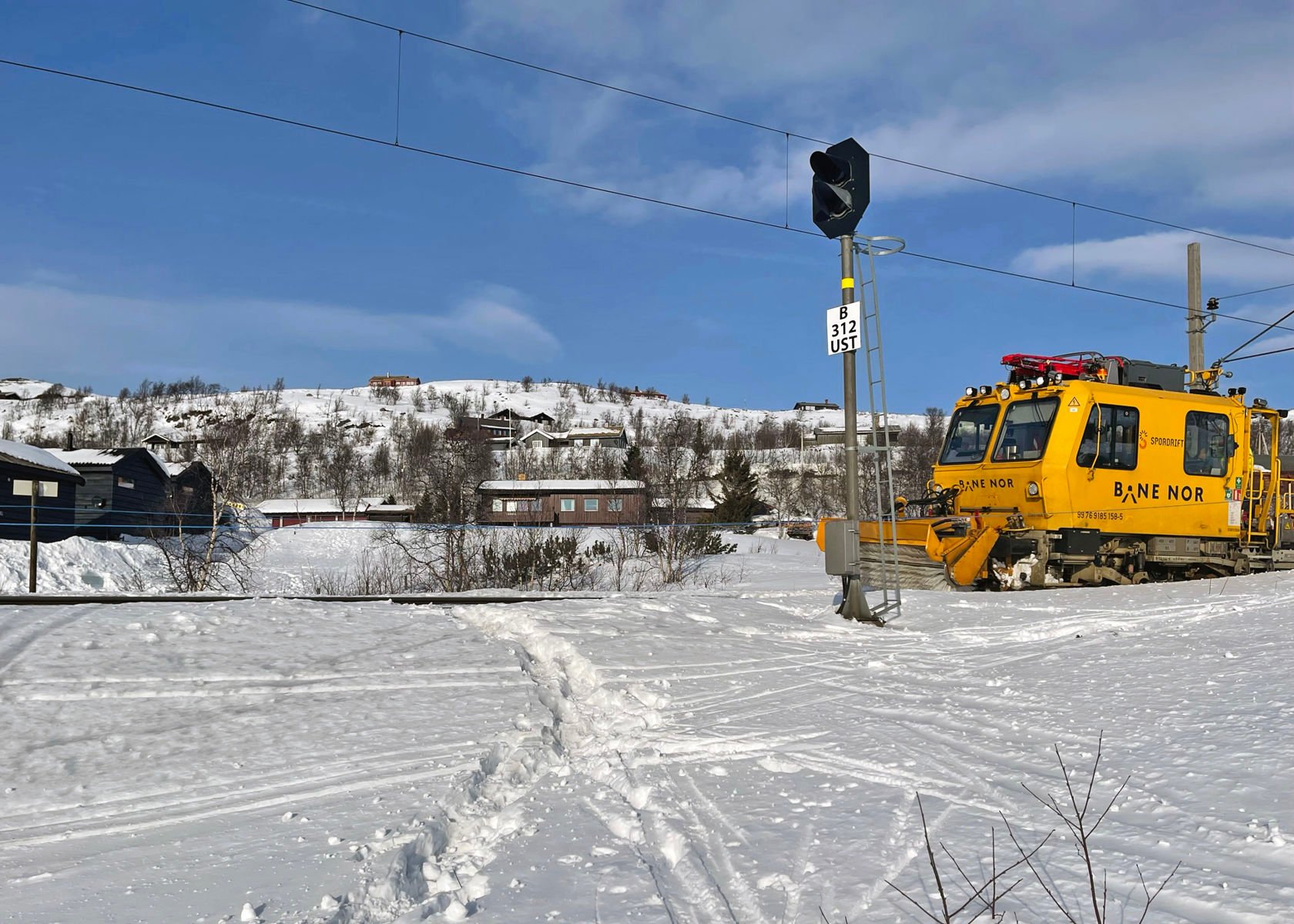 Spor i snøen krysser jernbaneskinnene. En gul arbeidsmaskin fra Bane NOR står på sporet.