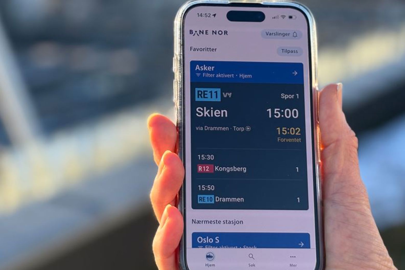 En hånd holder en mobil som viser avganger i NÅ-appen.