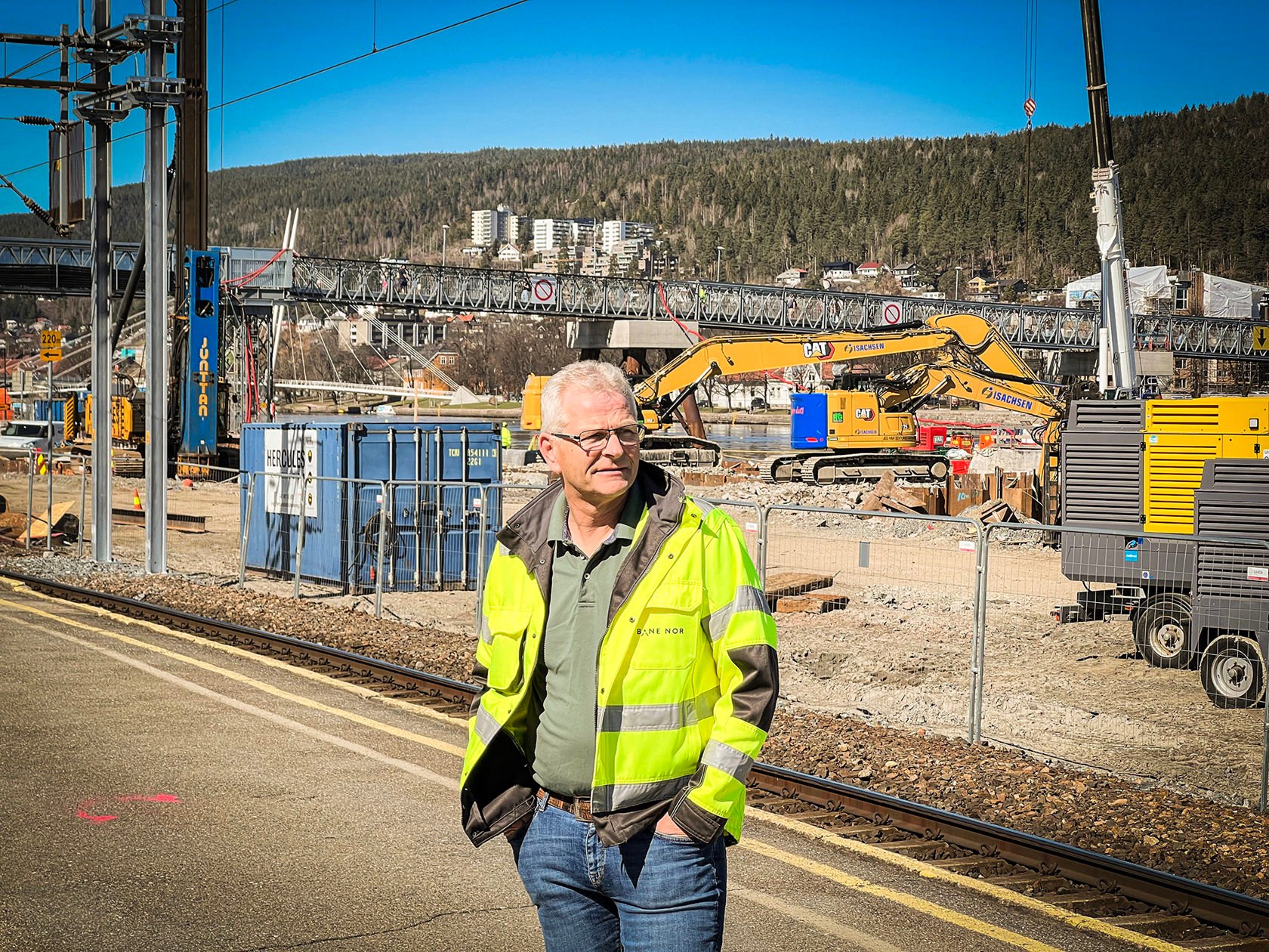 Mann i gul vernejakke står på plattform og ser mot høyre. I bakgrunn er anleggsområdet med maskiner.