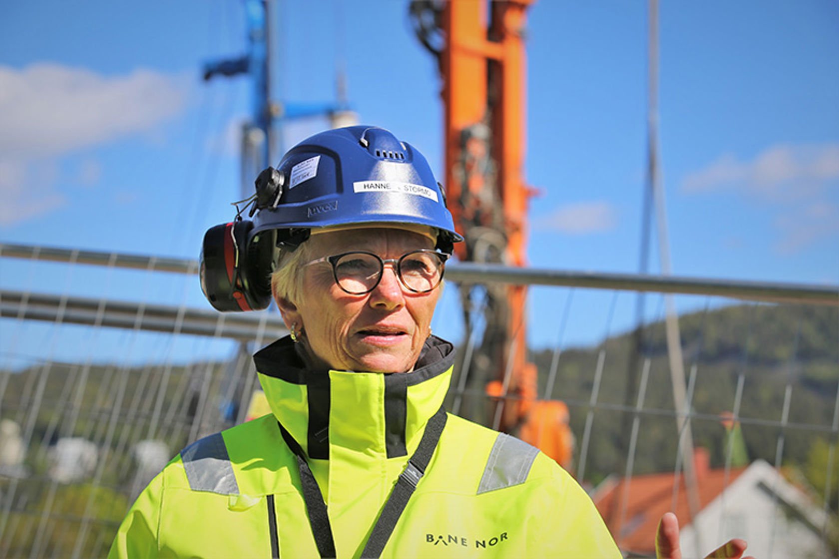 Kvinne i vernetøy med alvorlig mine står foran et flettverksgjerde med anleggmaskiner i bakgrunnen.