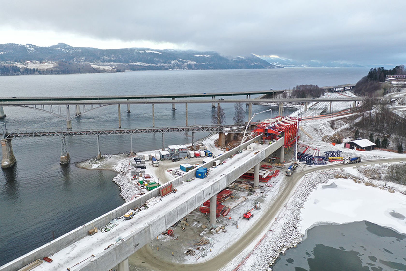 Dronebilde av brua, forskalingsvogna og betongbiler/pumper mens støpen pågår. I bakgrunnen Mjøsa og de andre bruene på Minnesund.