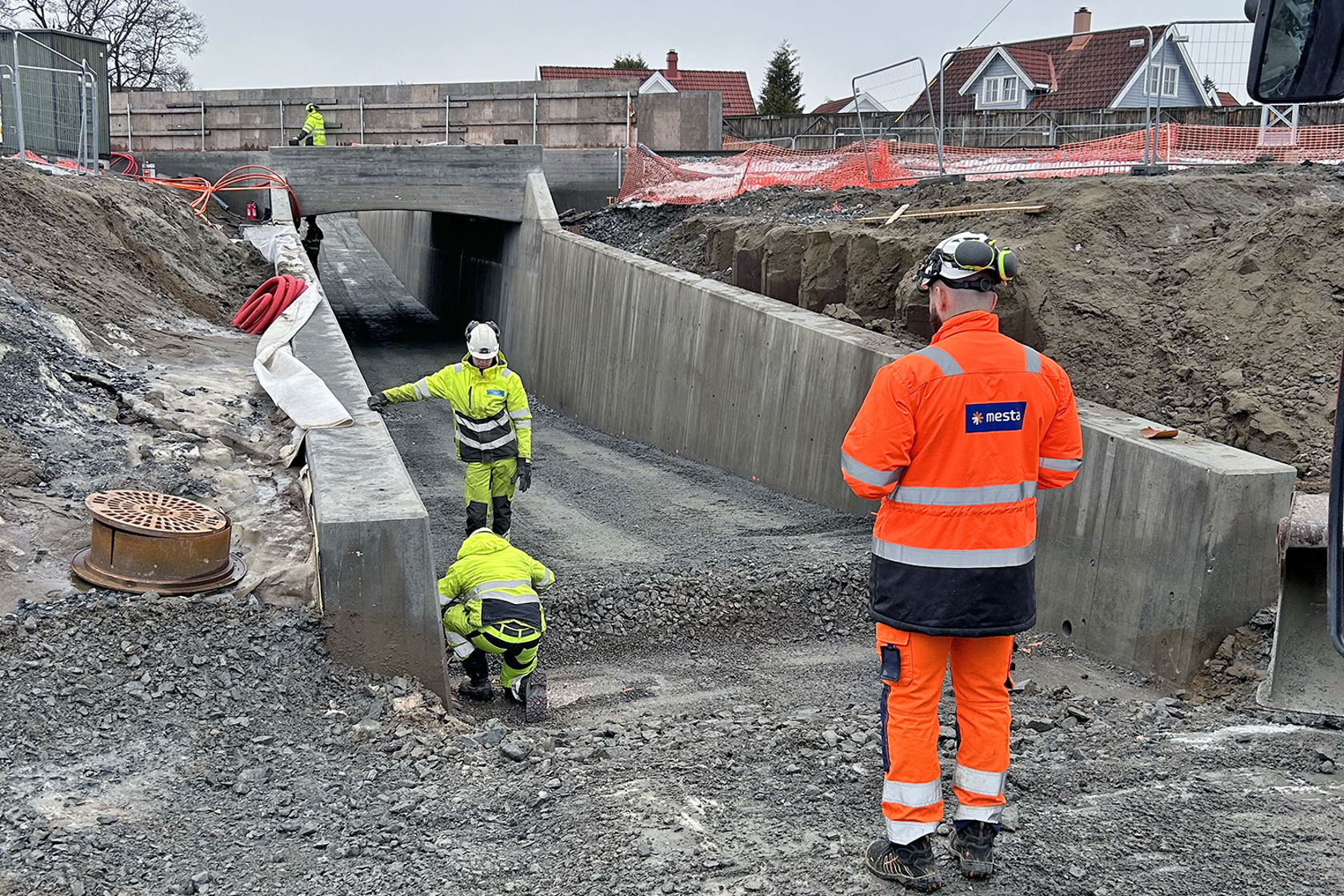 Arbeidere i anleggsklær i sving i undergangen, en gravemaskin skimtes i bildekanten.