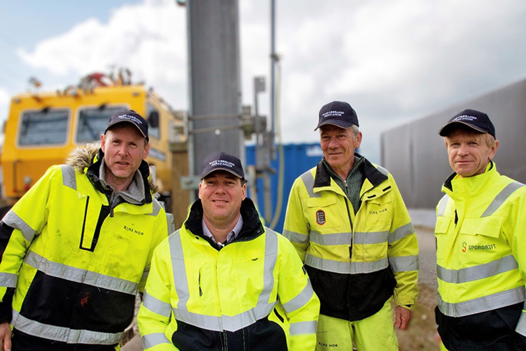 Ansatte på jernbanen i vernetøy og med caps på hodet, foran en stor gul maskin.