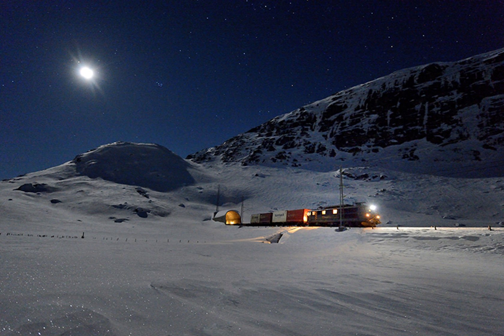 Et godstog kjører ut av en tunnel, med snøkledte fjell og en stjernehimmel.