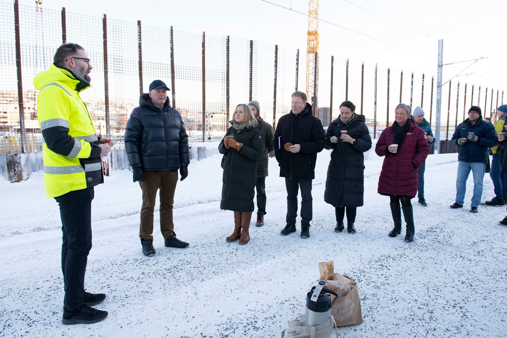 Gruppe mennesker med kaffe, gjerde og byggekran, vinter.