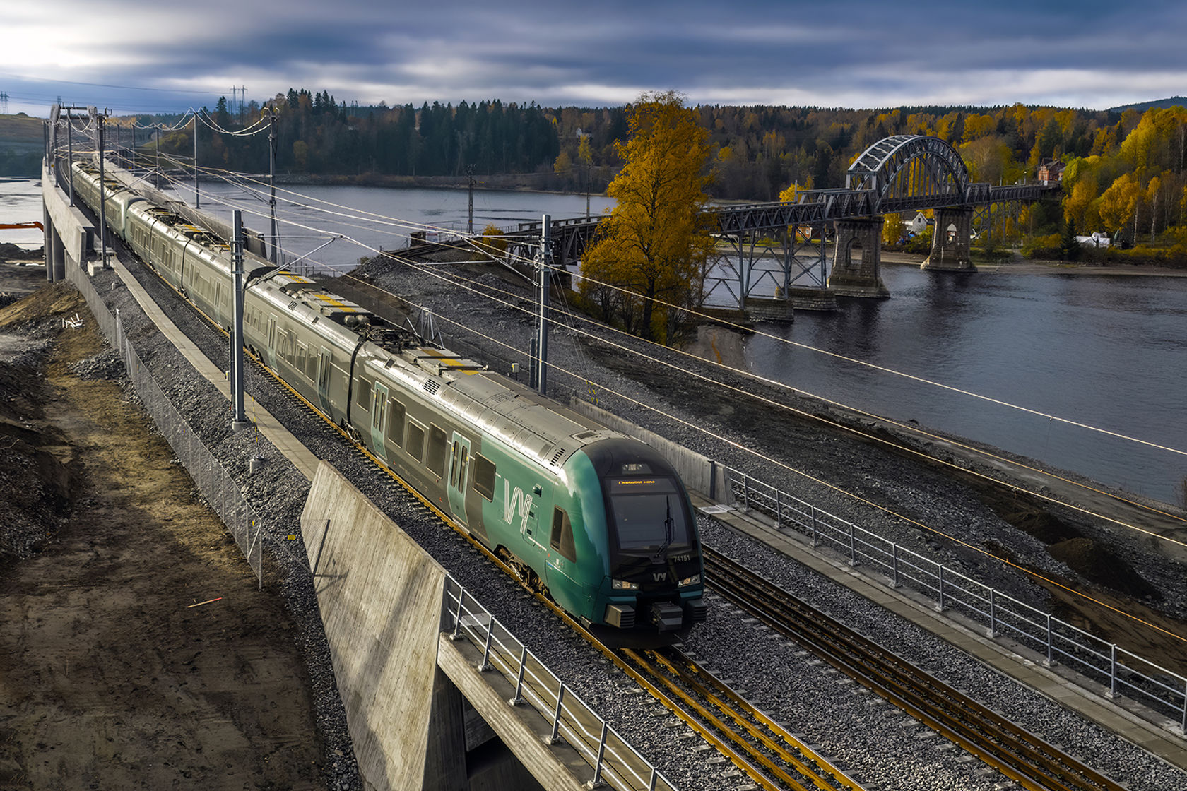 Et grønt Vy-tog med Vorma, gammel og ny jernbanebru i bakgrunnen.
