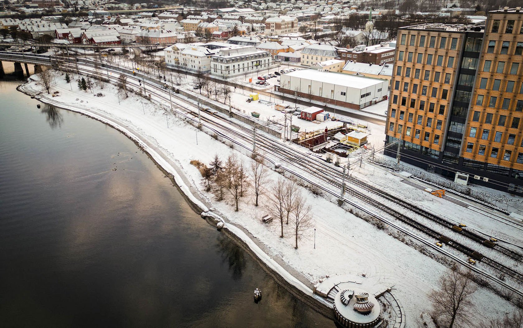 Sporområde og elvepromeade sett fra luften. Snø på bakken. Synlig anleggsområdet langs sporene.