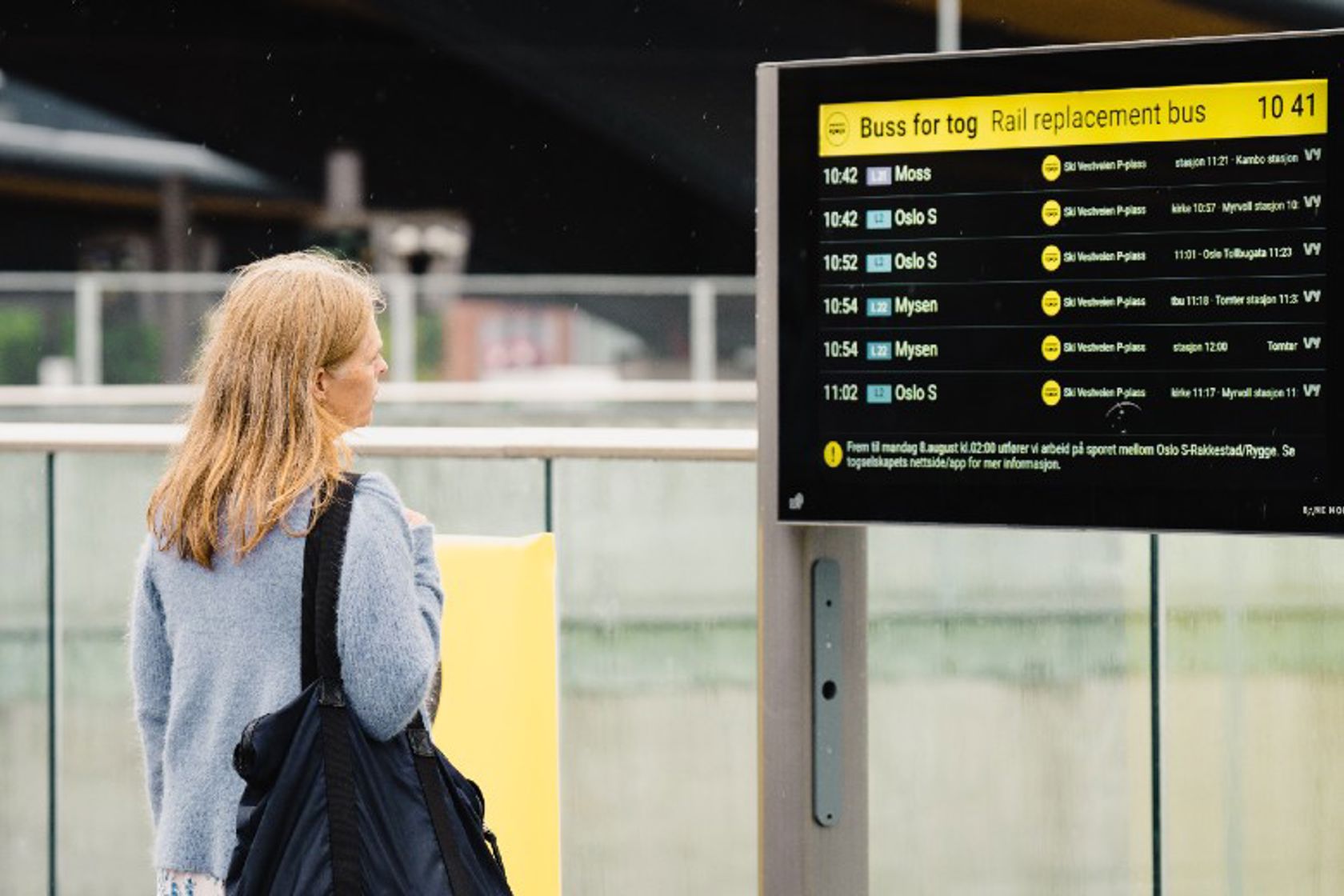 En kvinne som står å ser på en skjerm med informasjon og tider for buss for tog.