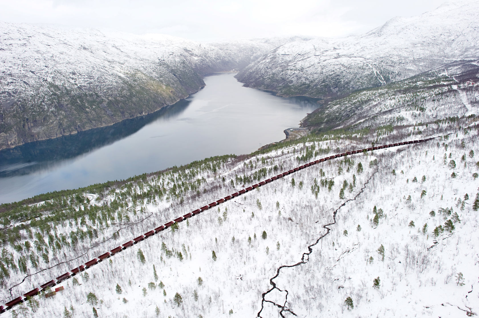 Bilde tatt fra lufta av et langt malmtog som går gjennom et hvit fjellandsskap med en innsjø.